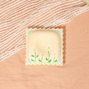 May include: A square, yellow ceramic dish with a scalloped edge and a light blue border. The dish is decorated with white daisies and green stems. The background is a peach-colored fabric.