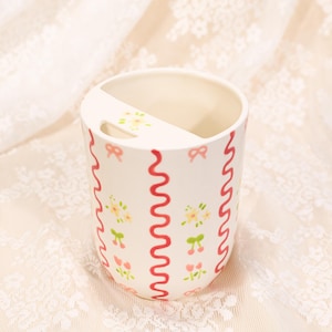 May include: A white ceramic toothbrush holder with a decorative design. The holder features red wavy lines, pink bows, and small floral and cherry patterns. The holder has a built-in divider.