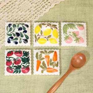 Könnte beinhalten: Fünf quadratische Keramikschalen mit gewelltem Rand, jede mit einem einzigartigen handbemalten botanischen Motiv: dunkle Oliven, gelbe Zitronen, rosa Erdbeeren, rote Tomaten und orangefarbene Karotten, dazu ein Holzlöffel.