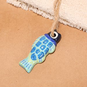 May include: A ceramic fish-shaped ornament with a blue and turquoise color scheme. The fish has a dark blue head and a light blue body with a scale pattern. A natural fiber cord is attached for hanging.