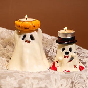 May include: Two Halloween-themed candle holders. One is a ghost with a pumpkin-shaped candle holder on its head. The other ghost wears a black hat and holds a knife. Both have lit candles.