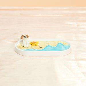 May include: A white ceramic tray with a beach scene featuring a small dog figurine, a yellow starfish, and painted blue water. The tray has a sandy yellow base and the word "Lola" is written on the sand.