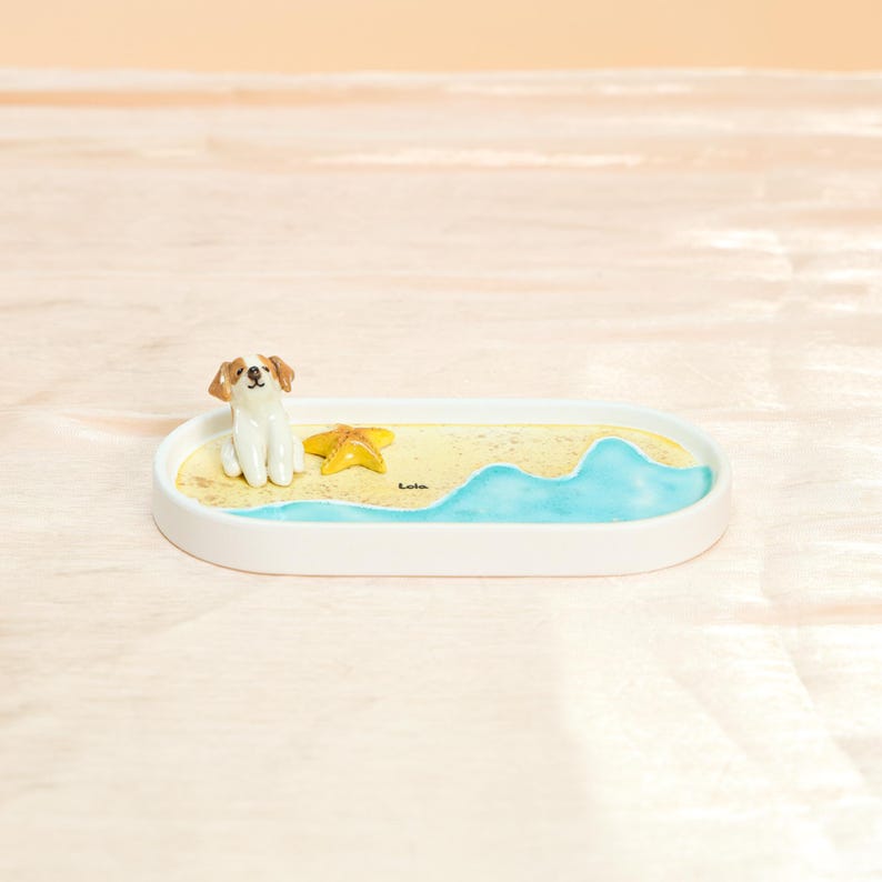May include: A white ceramic tray with a beach scene featuring a small dog figurine, a yellow starfish, and painted blue water. The tray has a sandy yellow base and the word "Lola" is written on the sand.