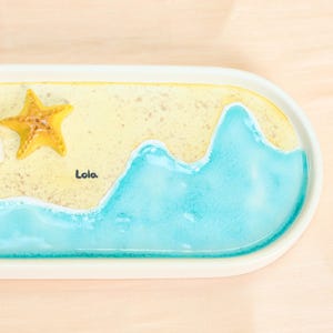 May include: A serving tray with a beach design. It features a yellow starfish, a sandy beach area, and a blue ocean wave design. The name "Lola" is written on the tray.