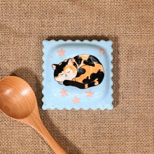 May include: A light blue, square ceramic dish with a scalloped edge. The dish features a sleeping calico cat illustration and pink flower accents. A wooden spoon is in the foreground.