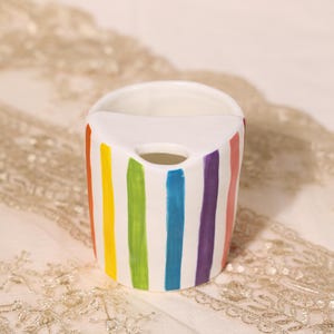 Pode incluir: Um porta-escova de dentes de cerâmica branca com listras verticais coloridas do arco-íris. O suporte tem um topo curvo e um orifício circular para uma escova de dentes. As listras incluem vermelho, laranja, amarelo, verde, azul, roxo e rosa.