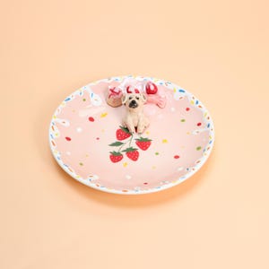May include: A pink ceramic plate with a playful design. It features a small, tan dog figurine, red strawberries, and colourful sprinkles. The rim is decorated with a white and blue pattern, and there are candies on the plate.