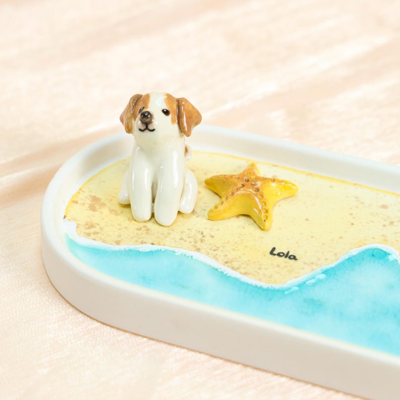 May include: A ceramic decorative tray with a painted beach scene, featuring a small dog figurine with brown and white markings, a yellow starfish, and blue water. The name "Lola" is written on the tray.