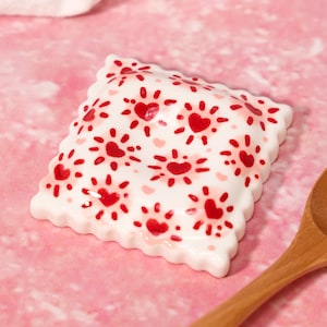 Puede incluir: Un plato cuadrado de cerámica con borde festoneado, que presenta una base blanca decorada con un patrón repetitivo de corazones rojos rodeados por líneas rojas radiantes. El plato tiene una forma ligeramente abombada, similar a un cojín, con un acabado brillante.