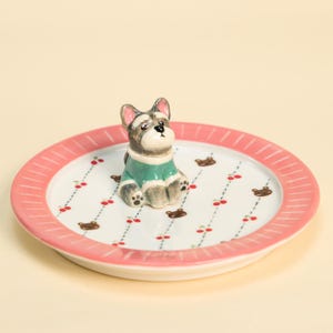 May include: A decorative ceramic plate with a pink rim and a white centre. The centre has a pattern of cherries and bear illustrations. A small, grey and white dog figurine wearing a green jumper sits on the plate.
