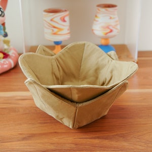 Puede incluir: Un bol de tela beige con un diseño plegado. El bol está sobre una superficie de madera.