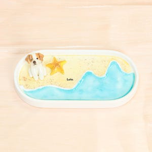 May include: An oval white decorative tray with a beach scene. It features a small dog figurine, a yellow starfish, and a blue wave design. The word "Lola" is written on the sand. The tray is set against a light wood background.