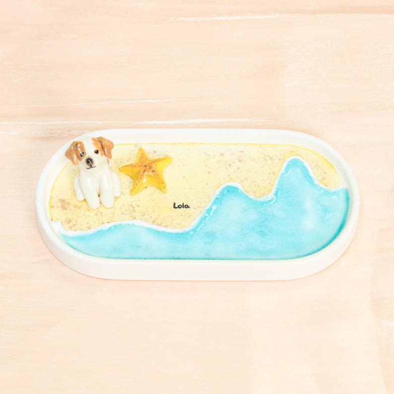 May include: An oval white decorative tray with a beach scene. It features a small dog figurine, a yellow starfish, and a blue wave design. The word "Lola" is written on the sand. The tray is set against a light wood background.