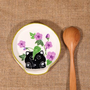 May include: A white ceramic spoon rest with a green rim, featuring a black cat design with purple flowers and green leaves. A wooden spoon rests beside the spoon rest on a burlap surface.