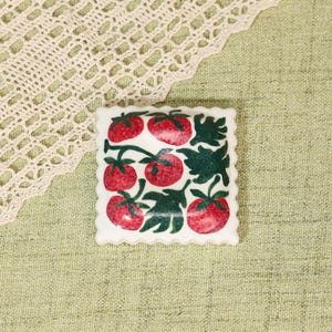 May include: A square, white ceramic tile with a scalloped edge, featuring a hand-painted design of red tomatoes and green leaves. The tomatoes are arranged in a repeating pattern, creating a charming, rustic aesthetic.