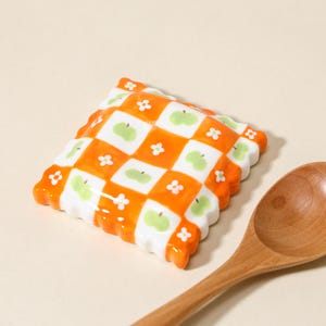 May include: A ceramic spoon rest with a checkered orange and white pattern, featuring green apple and white flower designs. The spoon rest has a scalloped edge. A wooden spoon is in the foreground.