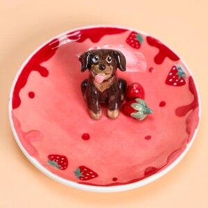 May include: A ceramic plate with a pink, strawberry-themed design. The plate features a brown dog figurine with its tongue out, along with painted strawberries. The rim is red with white accents.