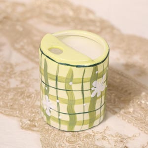 May include: A cylindrical ceramic container with a light yellow base, adorned with a green plaid pattern and white floral accents. The top features a curved, raised section with a small opening. The container is likely for holding small items.