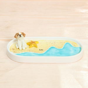 May include: An oval white decorative tray with a beach scene. It features a small dog figurine, a yellow starfish, and a painted ocean wave. The word "Lola" is written on the sand.