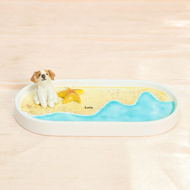 May include: An oval white decorative tray with a beach scene. It features a small dog figurine, a yellow starfish, and a painted ocean wave. The word "Lola" is written on the sand.