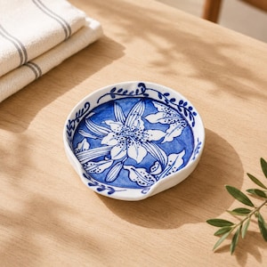 May include: A round ceramic spoon rest featuring a white base with a hand-painted blue floral pattern. The design displays a central lily-like flower surrounded by leaves and vines, with a scalloped edge and a small indentation for a spoon handle.