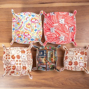 Peut inclure: Cinq plateaux de rangement en tissu avec différents motifs. Les plateaux sont beige, blanc et rouge avec divers motifs, notamment des chats, de la nourriture et des fleurs. Les plateaux ont une forme carrée avec des coins arrondis et sont faits de tissu avec une texture matelassée.