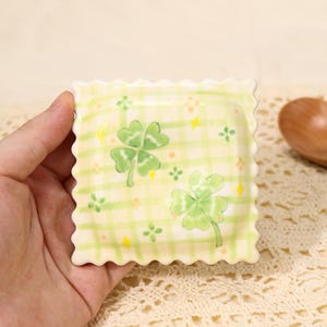 May include: A small, square ceramic dish with a scalloped edge. The dish features a light yellow plaid pattern with green shamrocks and small orange and yellow accents. The dish is held in a person's hand.