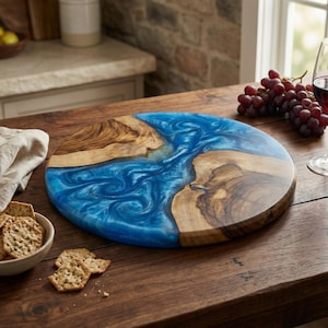Puede incluir: Una tabla redonda de madera para servir con un llamativo diseño de resina azul. La tabla presenta un patrón de vetas de madera natural y una sección de resina azul vibrante y arremolinada, ideal para aperitivos.