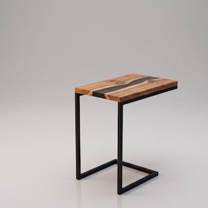 Puede incluir: Una mesa auxiliar rectangular con un tablero de madera natural que presenta una incrustación de resina oscura y ondulada en el centro. La mesa está sostenida por una estructura de metal negro en forma de C, que proporciona una base estable.