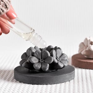 May include: A gray concrete flower diffuser with a small glass bottle of essential oil being poured onto the top. The diffuser is on a gray base and is sitting on a white surface.