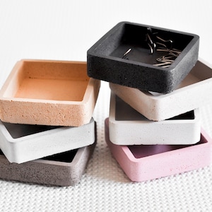 Könnte beinhalten: Ein Stapel kleiner, quadratischer Betonbehälter in verschiedenen Farben: Schwarz, Grau, Rosa und Beige. Die Behälter haben ein minimalistisches Design und sind übereinander gestapelt. Ein Behälter enthält silberne Büroklammern. Die Behälter stehen auf einer weißen, strukturierten Oberfläche.