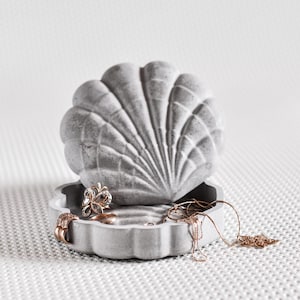 May include: A gray, seashell-shaped jewelry box with a textured surface. The box is open, revealing several pieces of jewelry, including rings and necklaces. The jewelry box is made of a solid material and sits on a white, textured surface.