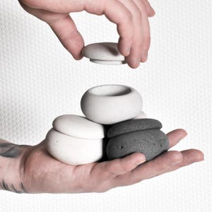 Puede incluir: Una mano sostiene una pila de pequeños objetos redondos, parecidos a piedras. Los objetos son en tonos de blanco y gris. Otra mano coloca una tapa encima de la pila. Los objetos parecen pequeños recipientes o piezas decorativas.