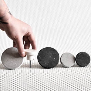 May include: Five round concrete knobs in various sizes, two are grey and three are black. The knobs are arranged in a row on a white textured surface.