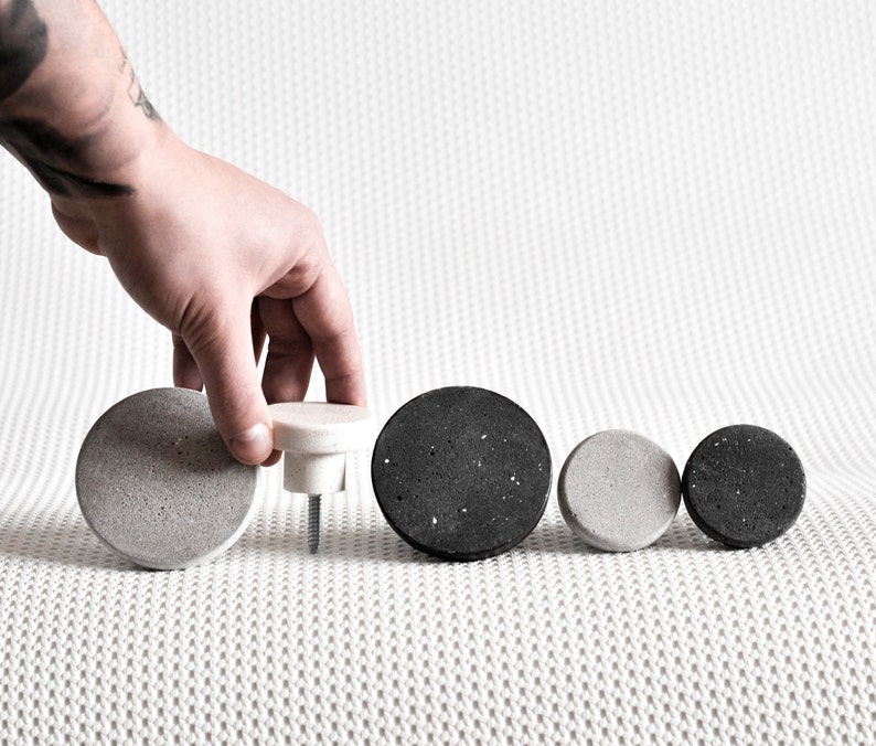 May include: Five round concrete knobs in various sizes, two are grey and three are black. The knobs are arranged in a row on a white textured surface.