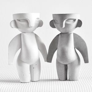 May include: Two white, stylized, concrete sculptures of human figures. The figures are standing side-by-side, facing forward, with their arms outstretched.