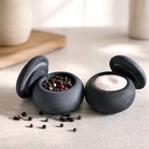 Puede incluir: Dos saleros y pimenteros de cerámica gris oscuro con tapas a juego. Un salero contiene sal, el otro granos de pimienta. Los saleros redondos están sobre una superficie clara. Un jarrón y otros artículos de cocina están en el fondo.