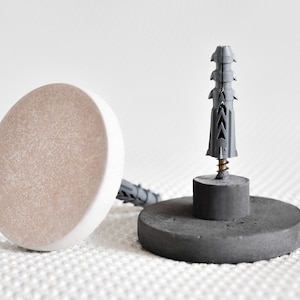 Puede incluir: Dos anclajes de pared de hormigón con un anclaje de pared de plástico gris en el centro. Los anclajes son redondos y tienen una parte superior plana. El anclaje de pared es de tamaño estándar y tiene un tornillo en el centro.