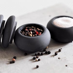 May include: A set of three dark gray, round, ceramic salt and pepper containers. One container holds white salt, another holds mixed peppercorns, and the third is a grinder. Scattered peppercorns are visible.