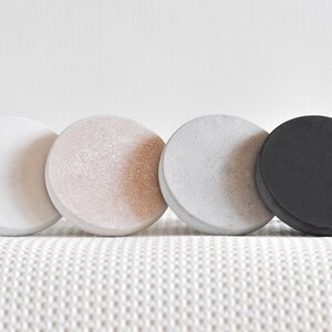 May include: Four round coasters in different colours: white, light pink, grey, and black. The coasters are made of a textured material and are arranged in a row.