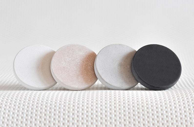 May include: Four round coasters in different colours: white, light pink, grey, and black. The coasters are made of a textured material and are arranged in a row.