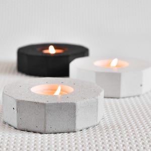 Pode incluir: Três castiçais geométricos de concreto cinza, branco e preto, cada um com uma vela acesa. As formas hexagonais têm um design moderno e minimalista, ideal para decoração de casa.