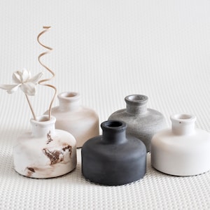 May include: A selection of small, round ceramic vases in neutral tones: white, grey, and black. One vase contains a white flower and a dried, curly stem. The vases are displayed on a textured white surface.