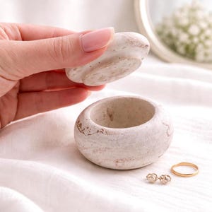 May include: A small, round, off-white stone trinket box with a lid being lifted. The box has a rough, textured surface with brown accents. Two small, flower-shaped earrings and a gold ring are next to the box. The background is a white, textured fabric.