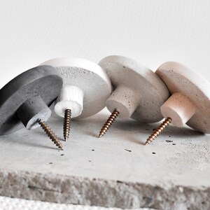 May include: Four concrete wall hooks in grey, white, and beige with copper screws. The hooks are arranged in a row on a grey concrete surface.