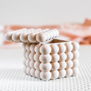 May include: A small, off-white decorative box with a lid, featuring a textured surface of rounded, bubble-like shapes. The lid is slightly ajar, revealing the interior. The box is set against a soft, neutral background.
