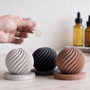 Może przedstawiać: Trzy sferyczne, teksturowane wazony dekoracyjne w kolorze szarym, czarnym i brązowym, każdy na pasującej okrągłej podstawie. Ręka trzyma zakraplacz nad wazonami. Butelki z brązowego szkła i gałązki lawendy w tle.