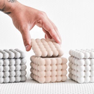 Könnte beinhalten: Drei quadratische dekorative Boxen mit abgerundeten, blasenartigen Texturen. Eine Box ist geöffnet und zeigt ihr Inneres. Die Boxen sind in Grautönen, Beige und Weiß gehalten. Eine Hand greift nach der offenen beigen Box.