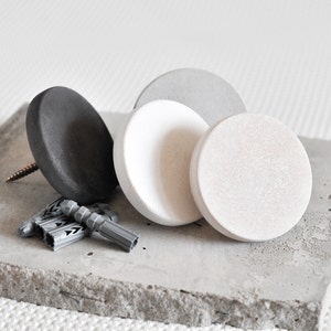 May include: Four round concrete wall hooks in grey, white and black. The hooks are on a grey concrete surface.