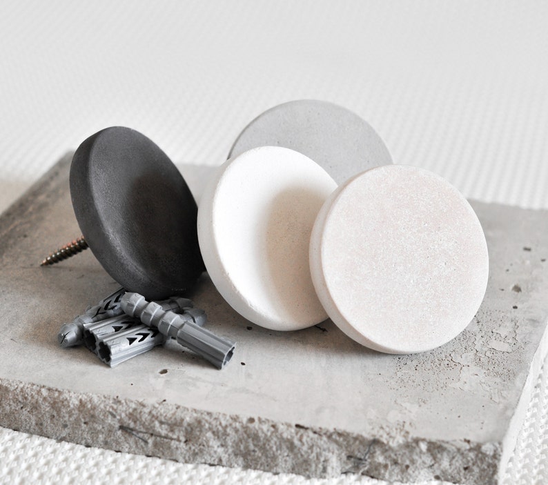 May include: Four round concrete wall hooks in grey, white and black. The hooks are on a grey concrete surface.